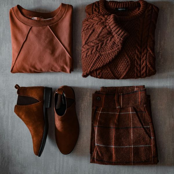 Flat-lay of a cozy fall outfit: rust sweatshirt, cable-knit sweater, suede ankle boots and plaid trousers
